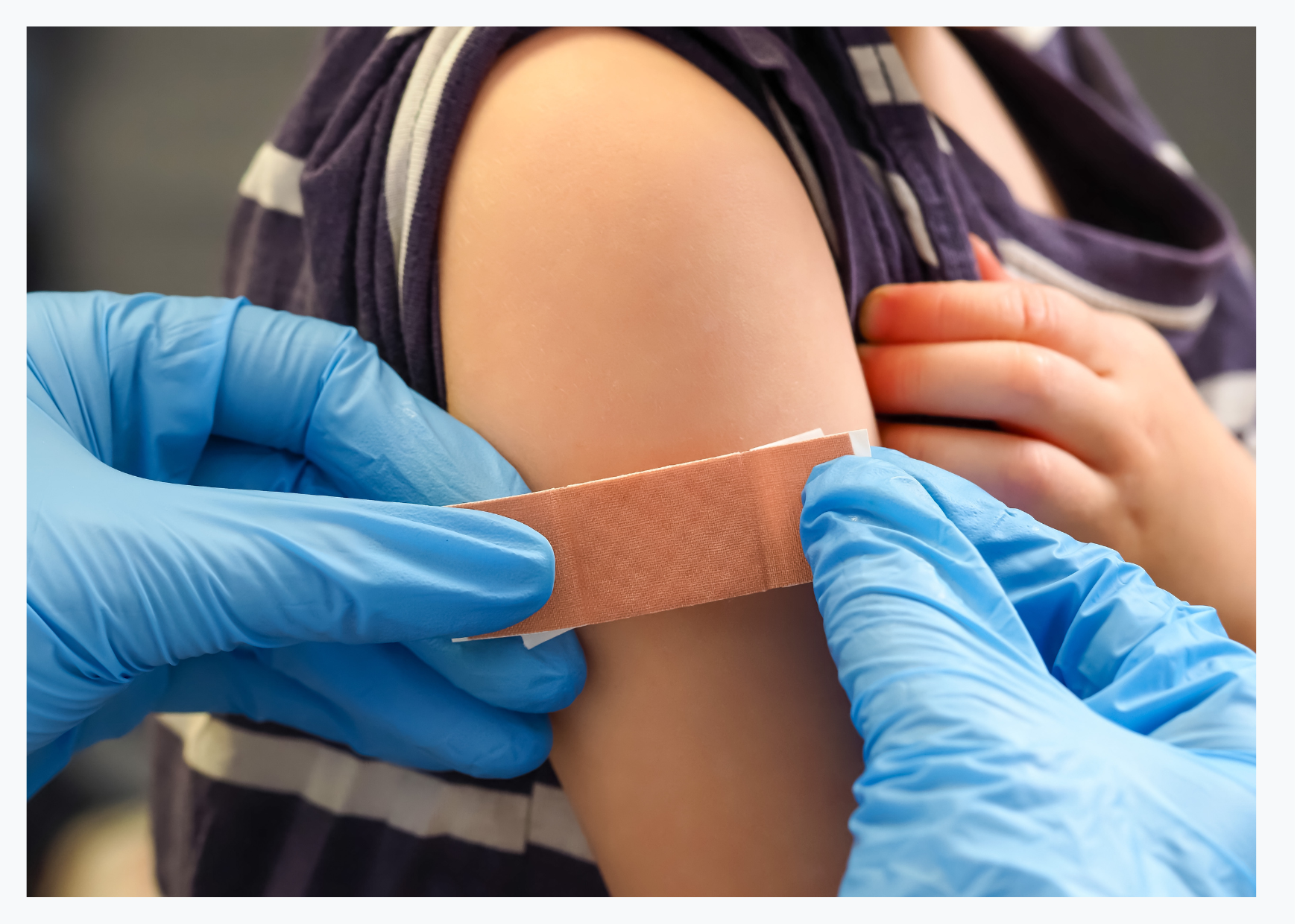 A close up the upper arm of a white child wearing a blue and white striped t-shirt. Adult hands wearing medical gloves can be seen applying plaster to the child's arm.