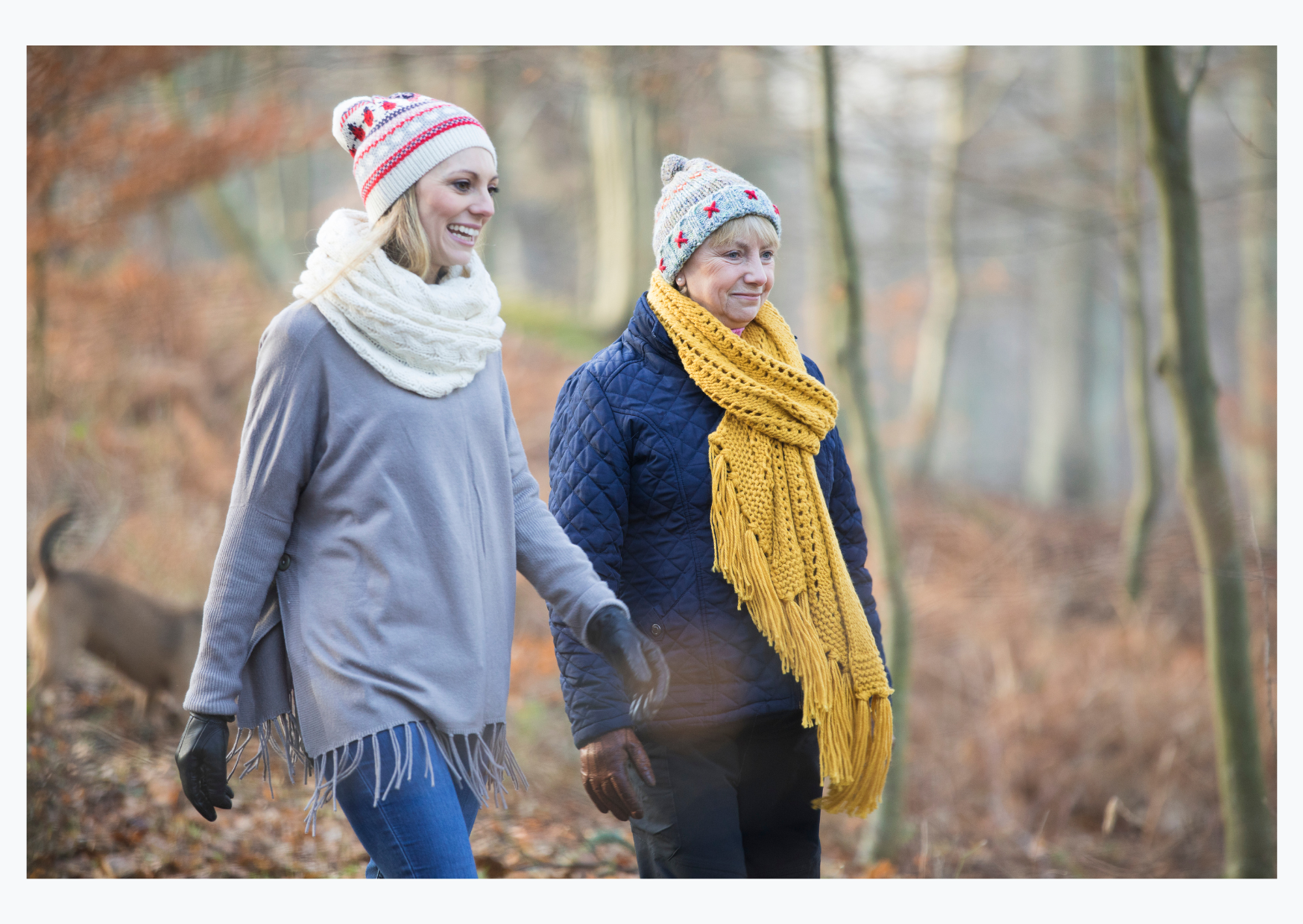 Two women in warm clothes and wool hats are walking in a wood and smiling.