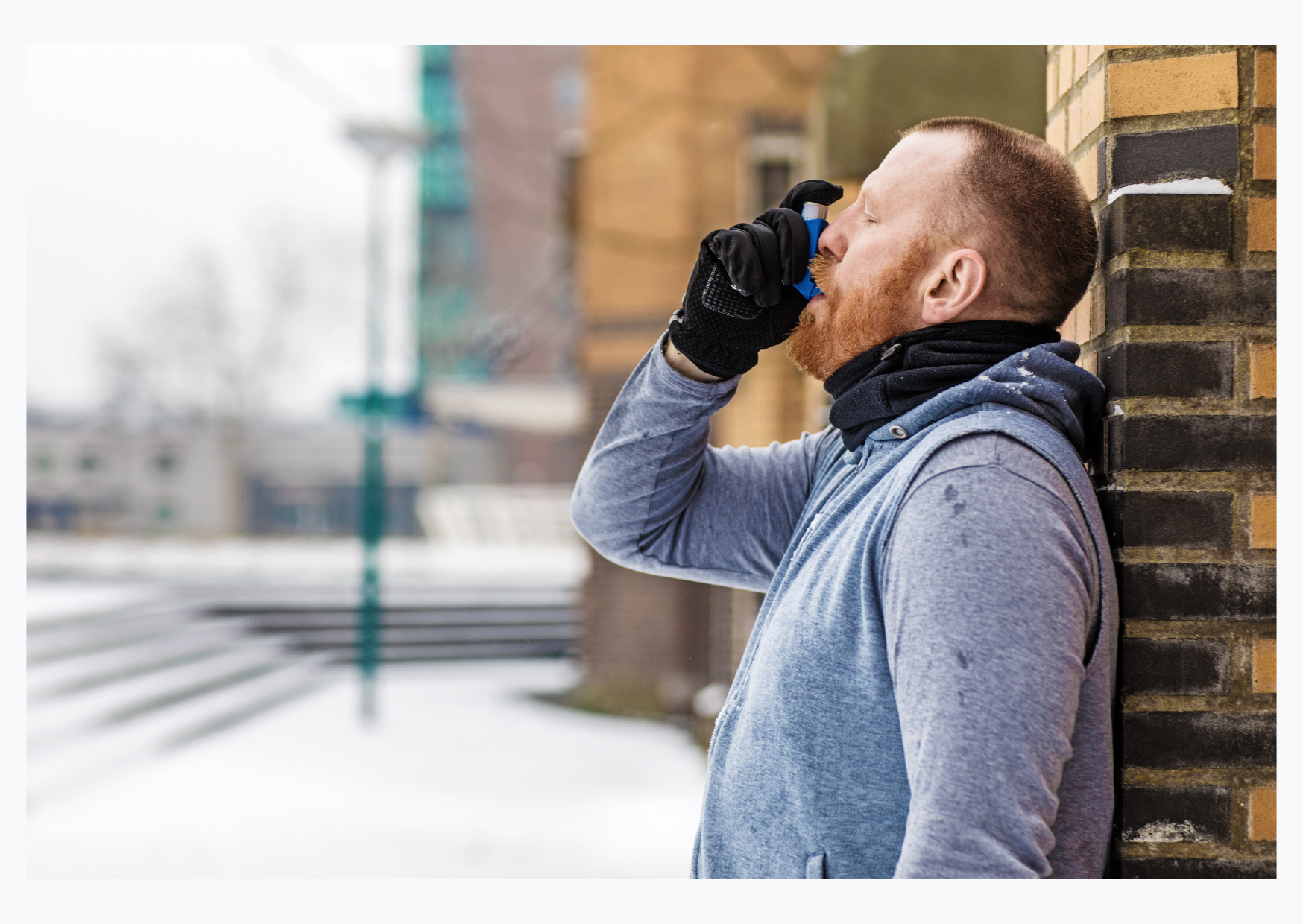 A white man with red hair is leaning against a wall using an asthma inhaler. He is dressed warmly in a grey jumper and black scarf and gloves.