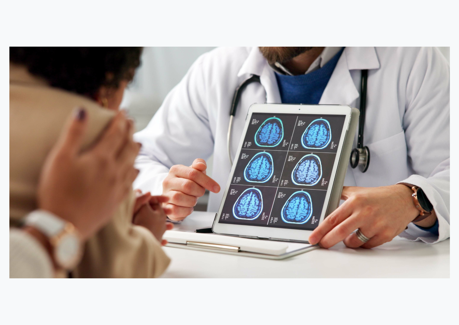 A doctor is holding images of a brain scan towards a patient being comforted by a loved one