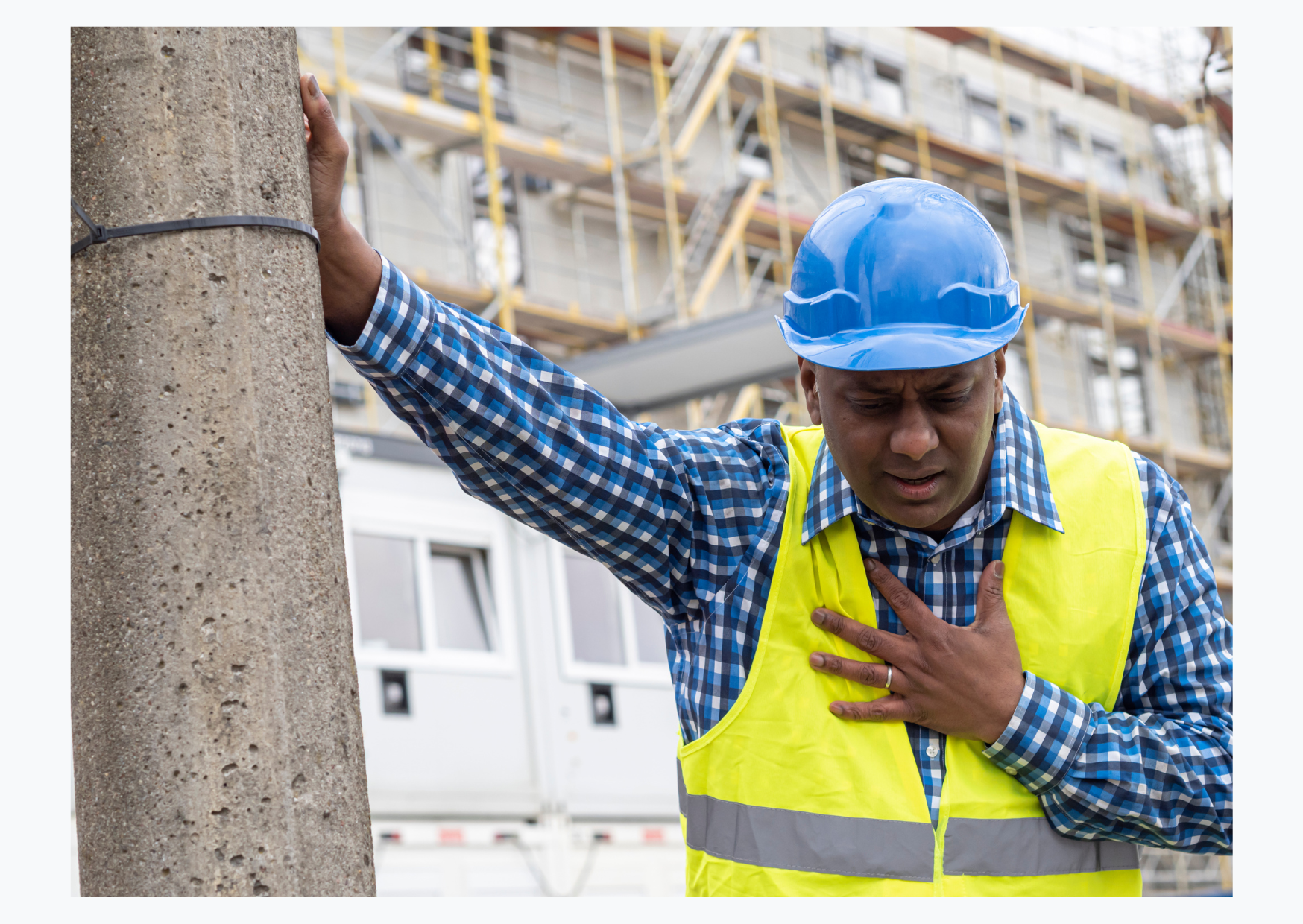A man on a construction site wearing a hard hat and hi-vis jacket leans on a post, clutching his chest