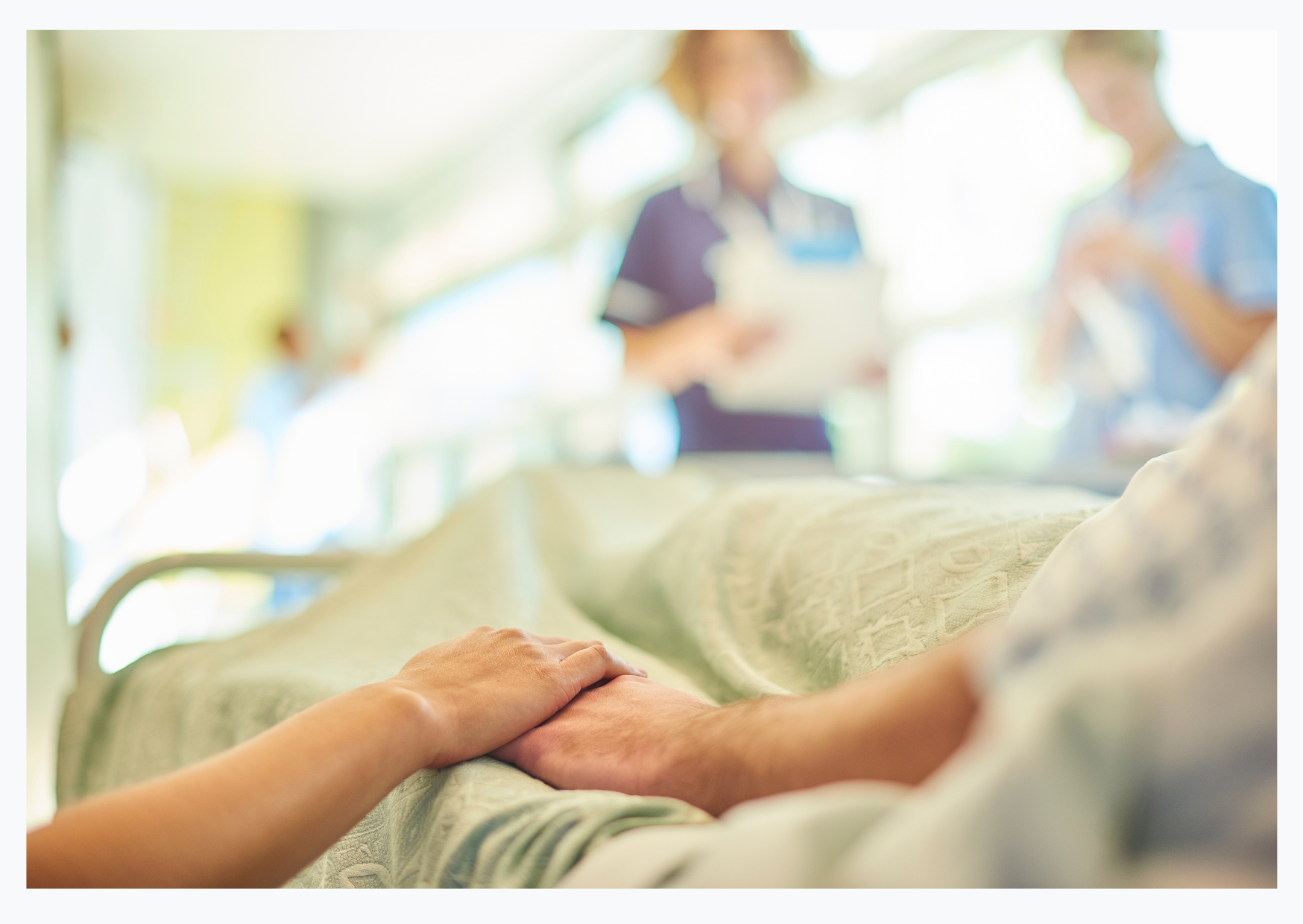 Two hands are clasping on a hospital bed at Norfolk and Norwich University Hospital, medical professionals can be seen in the background.